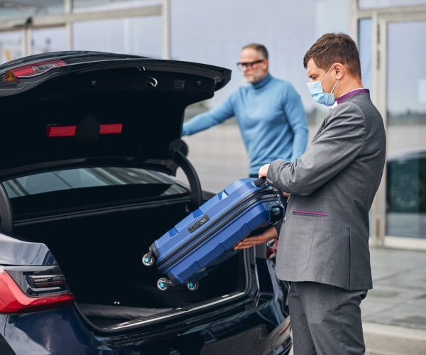 Serious elegant dark-haired Caucasian airport driver putting his client traveling bag in the car trunk