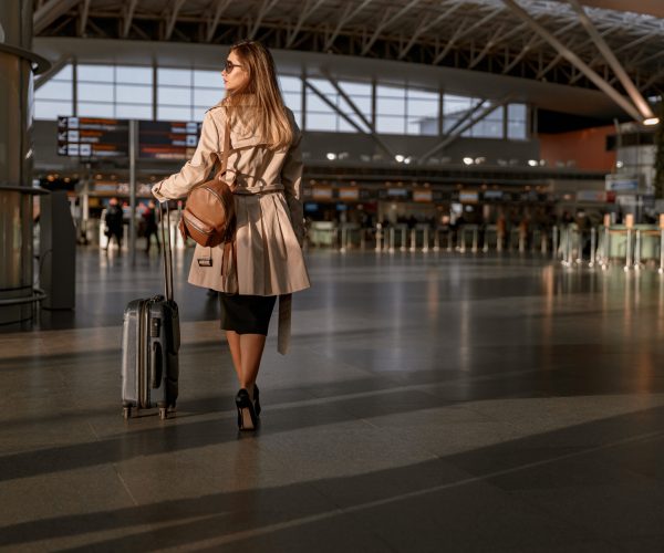 Back view of young woman holding suitcase while walking in arrival or departure hall in airport