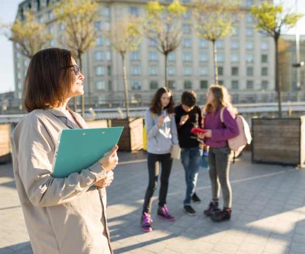 Portrait of mature smiling female teacher in glasses with clipboard, outdoor with a group of teenagers students, golden hour