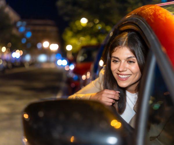 Young woman leaning out of a car window at night, smiling as city lights and bokeh blur by, enjoying a joyful, free evening ride through urban streets